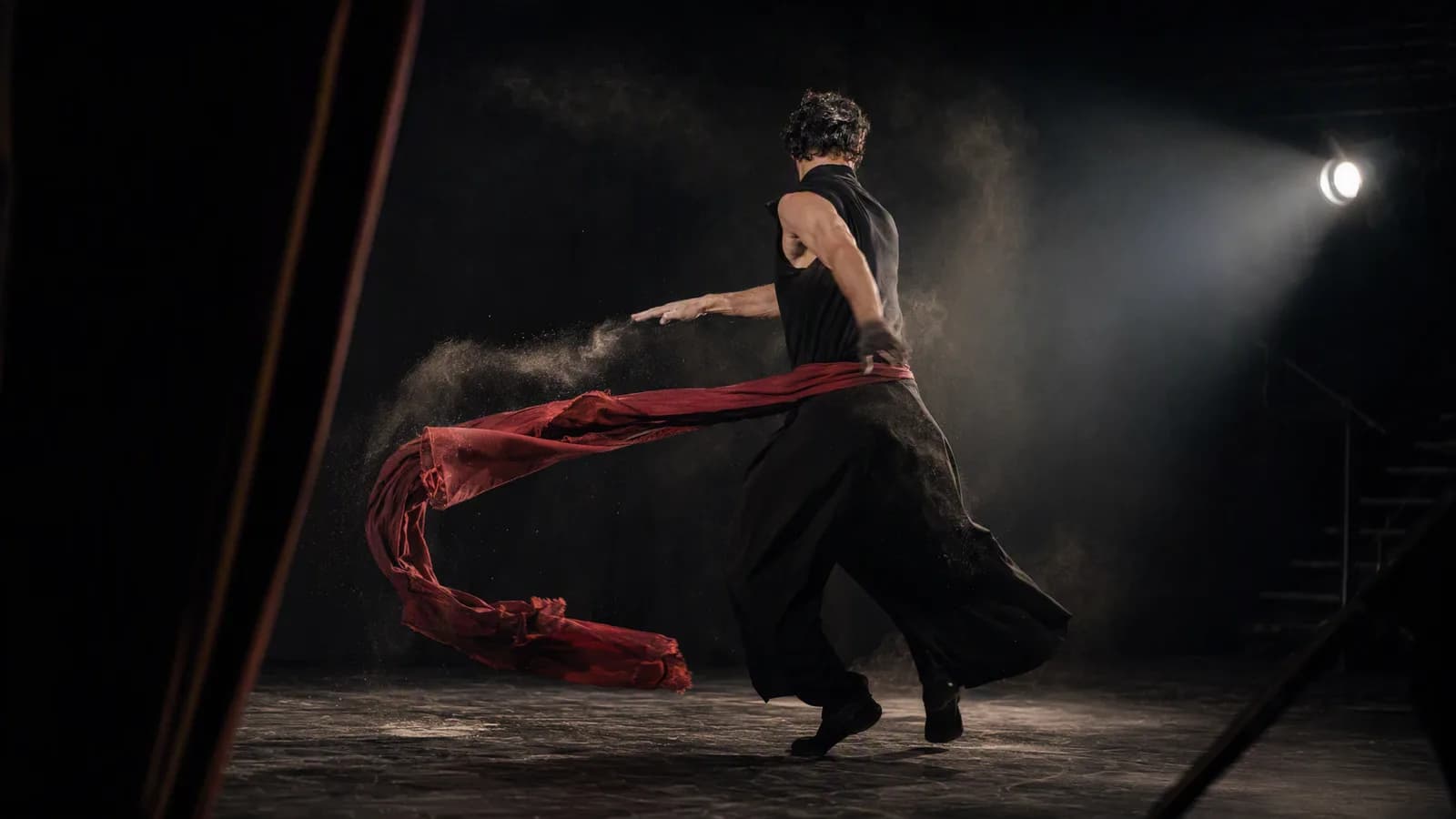 A dancer turning with red fabric and dust suspended in stage light